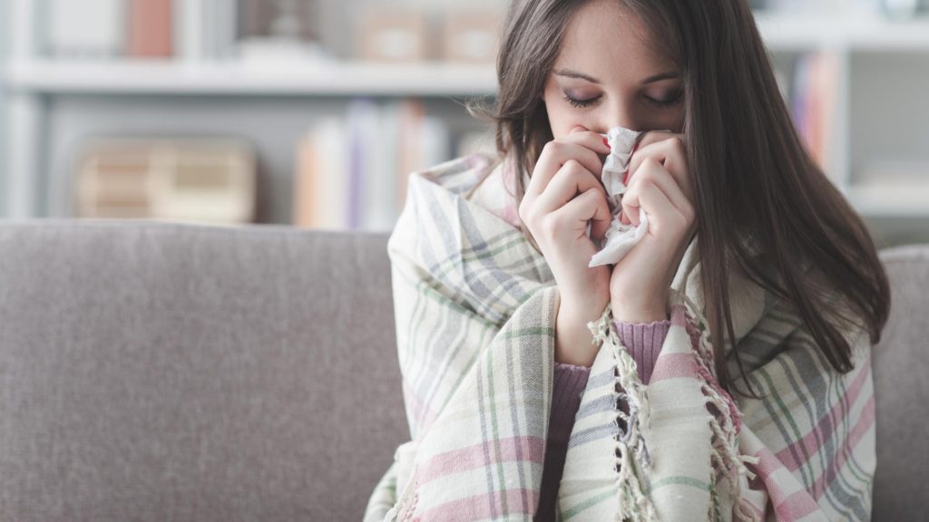 A young woman sitting on the couch, wrapped in a blanket, and blowing her nose