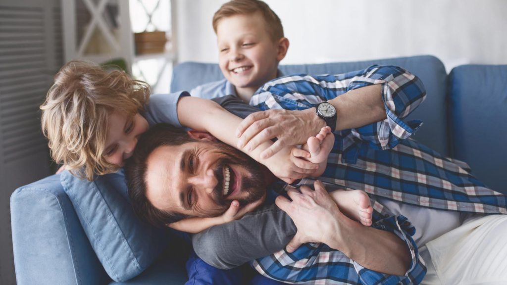 A father and his two sons laughing and playing on a couch