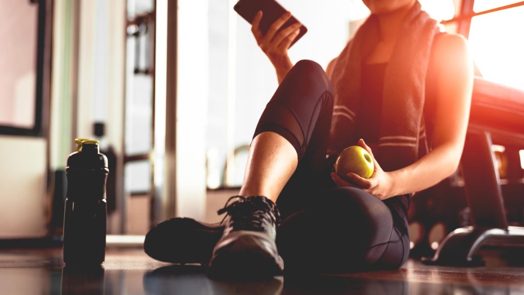Close up of woman using smart phone and holding apple while workout in fitness gym.
